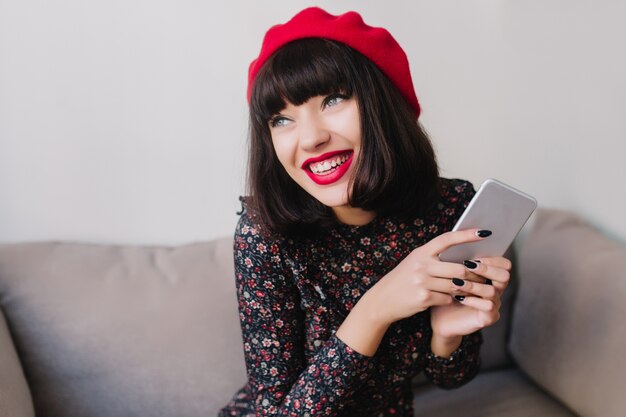 Chica morena alegre con vestimenta retro y boina roja esperando llamada de un amigo, sujetando smartphone plateado