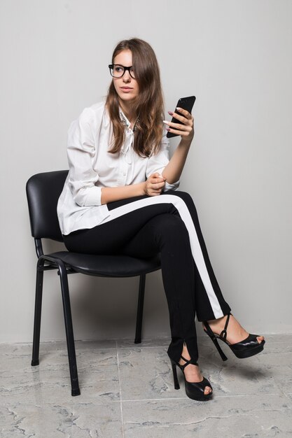 Chica morena alta con gafas vestidas con camiseta blanca y pantalón negro se sienta con el teléfono en la silla de oficina delante de un fondo blanco