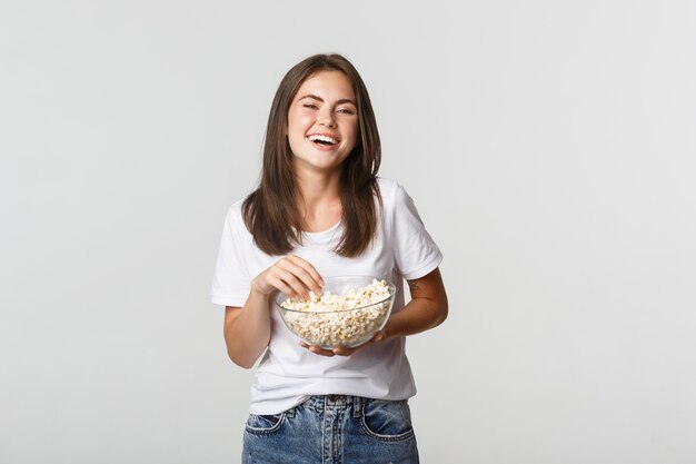 Chica morena atractiva alegre riéndose de la película de comedia, comiendo palomitas de maíz.