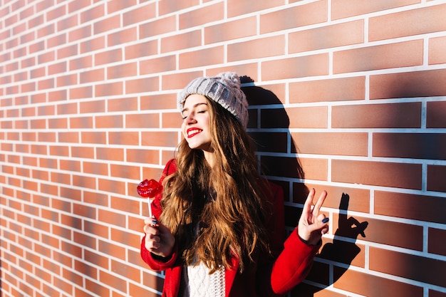 Chica morena atractiva con el pelo largo en abrigo rojo escalofriante en el sol en la pared exterior. Viste gorro de punto, tiene labios rojos de piruleta.