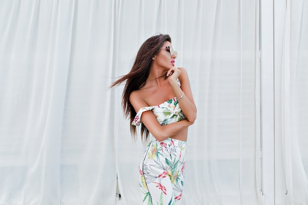 Chica morena atractiva con el pelo largo está posando junto a las largas cortinas blancas al aire libre. Ella está mirando lejos.