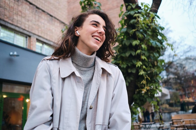 Chica morena bastante alegre en gabardina felizmente escuchando música en auriculares inalámbricos en un café en la calle de la ciudad