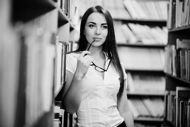 Chica morena en la biblioteca con gafas en la mano, blusa blanca y minifalda negra. Mujer de negocios sexy o concepto de maestro. Foto en blanco y negro.