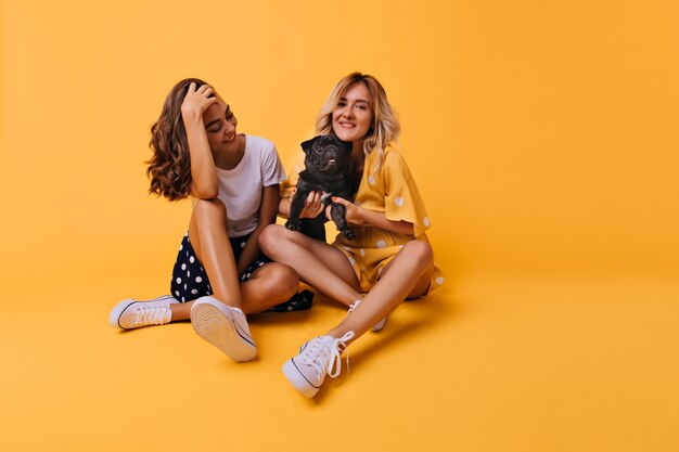 Chica morena bien vestida jugando con su cabello y mirando bulldog francés. Señora joven rubia magnífica que sostiene el perrito lindo.