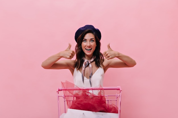 Foto gratuita la chica morena de boina azul está contenta con las compras y muestra los pulgares para arriba. mujer feliz de buen humor en poses de sombrero morado y blusa blanca sobre fondo rosa.