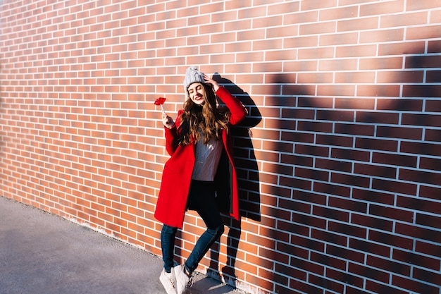 Chica morena bonita de longitud completa con el pelo largo en abrigo rojo escalofriante en el sol en la pared exterior. Lleva un gorro de punto, sostiene los labios rojos como una piruleta, mantiene los ojos cerrados y sonríe.