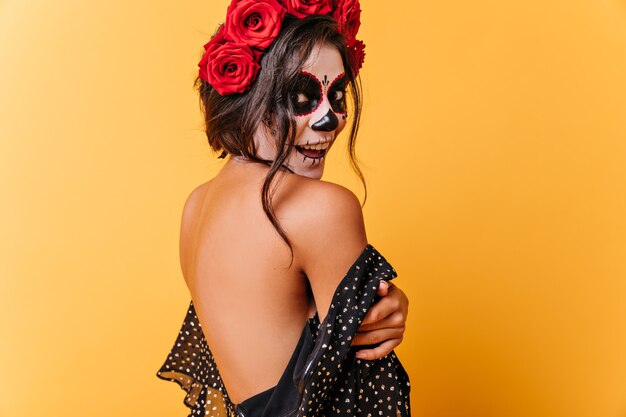 Chica morena bronceada posa felizmente en vestido con espalda abierta. Señora con maquillaje de Halloween sorprendida mirando a cámara.