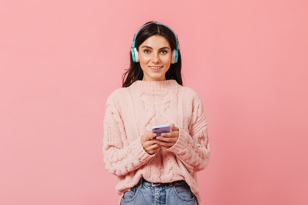 Chica morena bronceada con sonrisa encantadora mira a la cámara. Señora en auriculares cambia la canción en el teléfono.