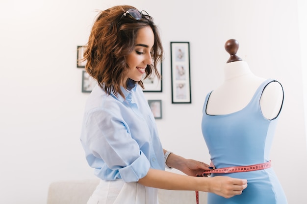 Una chica morena con una camisa azul está trabajando en el estudio del taller. Ella se adapta al vestido azul en el maniquí.