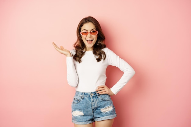 Foto gratuita chica morena coqueta y elegante, riendo y sonriendo, con gafas de sol, blusa blanca y jeans, fondo rosa. copia espacio