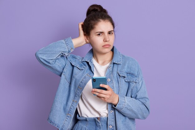 Chica morena desconcertada vistiendo chaqueta vaquera y camisa blanca, manteniendo la mano en el dorso de la mano, sosteniendo un teléfono inteligente