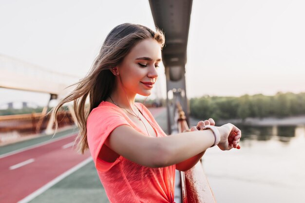 Chica morena despreocupada mirando el reloj inteligente con una sonrisa suave Retrato al aire libre de una adorable corredora usando una pulsera de fitness
