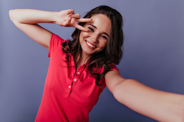 Chica morena dichosa en traje rojo jugando. Retrato de dama de buen humor haciendo selfie en pared púrpura.