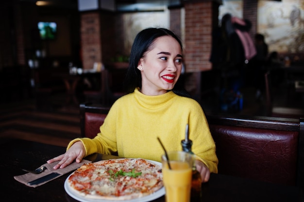 Chica morena divertida en suéter amarillo comiendo pizza en el restaurante