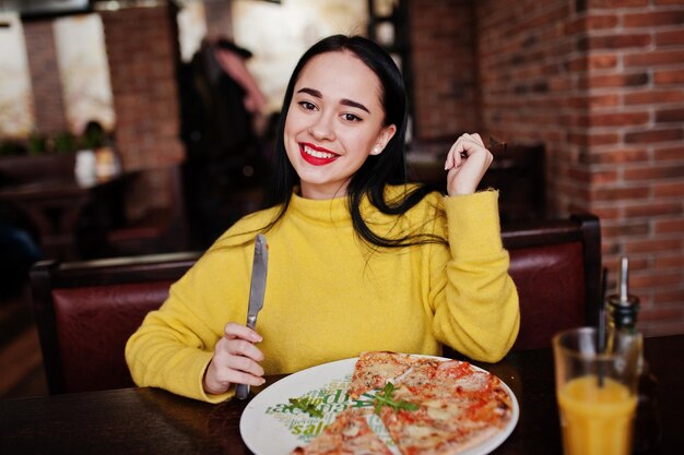 Chica morena divertida en suéter amarillo comiendo pizza en el restaurante