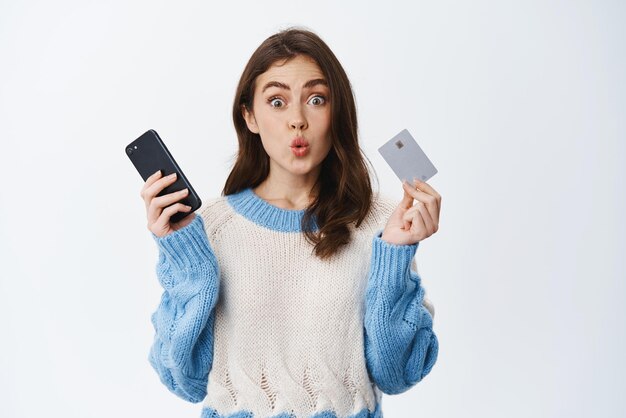 Una chica morena emocionada dice wow y se ve asombrada sosteniendo una tarjeta de crédito de plástico y comprando un teléfono inteligente en línea o usando la aplicación de banca móvil con fondo blanco