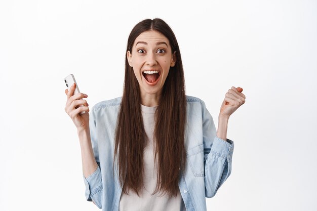 Chica morena emocionada grita asombrada, sosteniendo un teléfono inteligente y celebrando, ganando en línea, recibe buenas noticias, logra el objetivo de la aplicación, de pie sobre una pared blanca