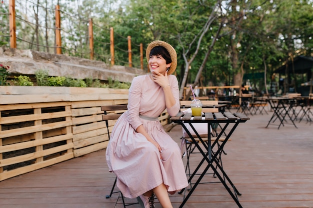 Chica morena emocionada en hermoso vestido antiguo tocando la cara con una sonrisa encantadora descansando en la cafetería al aire libre