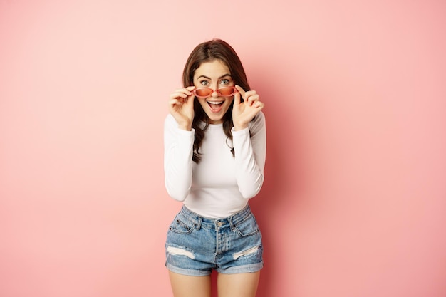 Chica morena entusiasta con elegantes gafas de sol, reacciona sorprendida y asombrada, mirando con incredulidad a la cámara, de pie sobre un fondo rosa