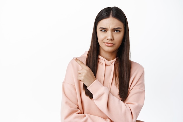 Chica morena escéptica, señalando con el dedo en el logo de la esquina superior izquierda, teniendo dudas, frunciendo el ceño y mirando insegura al frente, de pie contra la pared blanca