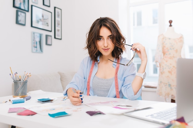 Una chica morena está sentada a la mesa en el taller. Ella tiene camisa azul y un desorden creativo en la mesa. Ella tiene gafas y un lápiz en las manos y mira a la cámara.