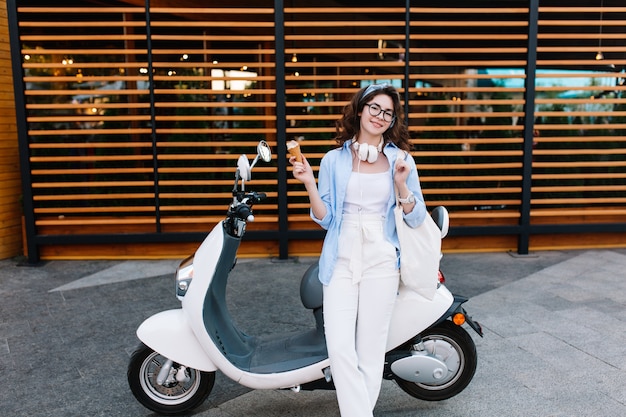 Chica morena con estilo en gafas y chaqueta azul comiendo helado después de ir de compras