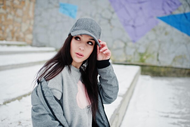 Chica morena con estilo en gorra gris estilo casual de la calle en el día de invierno contra la pared de color