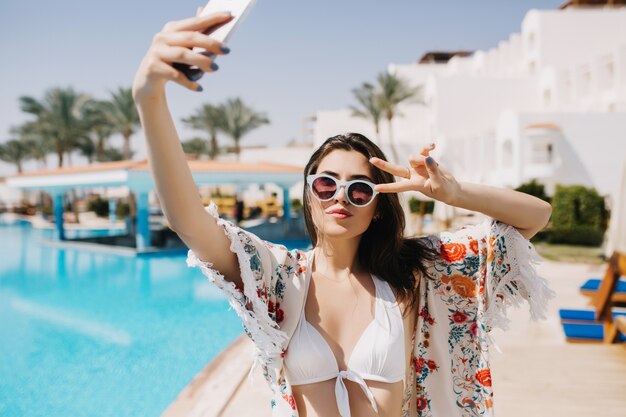 Chica morena con estilo guapa descansando en el resort del sur y haciendo selfie antes de la fiesta en la piscina con amigos. Retrato de hermosa mujer joven con gafas de sol y camisa de moda divirtiéndose en vacaciones