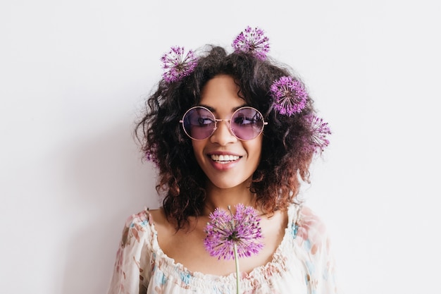 Chica morena feliz en gafas de moda posando con flores en el pelo. Mujer africana rizada con pie de allium púrpura.