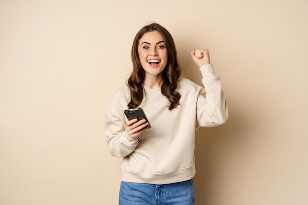 Chica morena feliz sosteniendo un teléfono inteligente y animando, ganando, celebrando la victoria en la aplicación de teléfono móvil, de pie sobre un fondo beige.