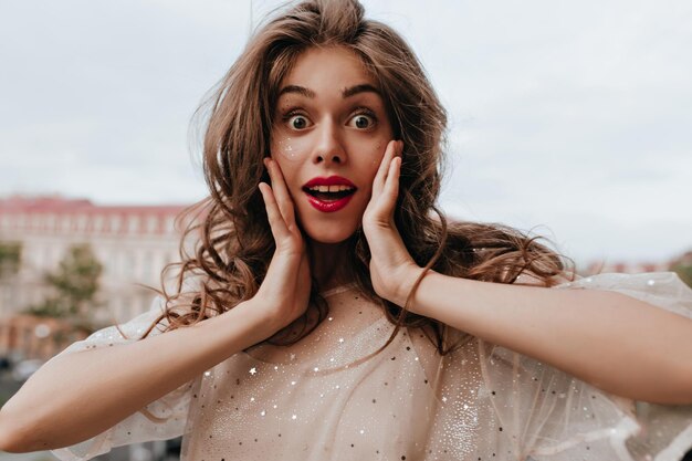 Foto gratuita chica morena feliz en vestido blanco mira a la cámara retrato de mujer feliz en traje con destellos posando en el balcón