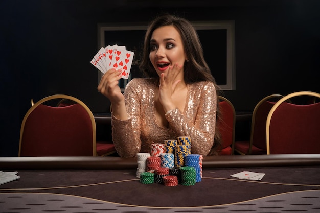 Una chica morena feliz con un vestido dorado brillante está jugando al póquer sentada en la mesa del casino. Ella está haciendo apuestas esperando una gran victoria y mostrando sus cartas. Juegos de azar por dinero. Juegos de fortuna.