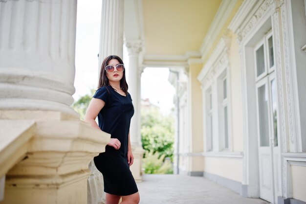 Chica morena con gafas de sol de vestido negro posadas contra la antigua casa vintage en la calle de la ciudad