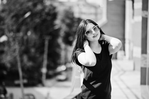 Chica morena con gafas de sol de vestido negro posando en la calle de la ciudad