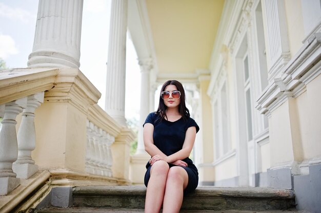 Chica morena con gafas de sol de vestido negro sentada en las escaleras de una antigua casa vintage posando en la calle de la ciudad