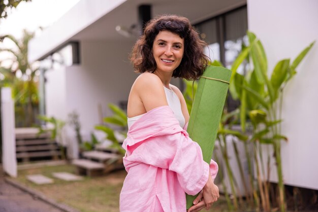 Chica morena joven caucásica feliz en camisa rosa sosteniendo estera de yoga sonriendo a la cámara al aire libre Concepto de estilo de vida saludable