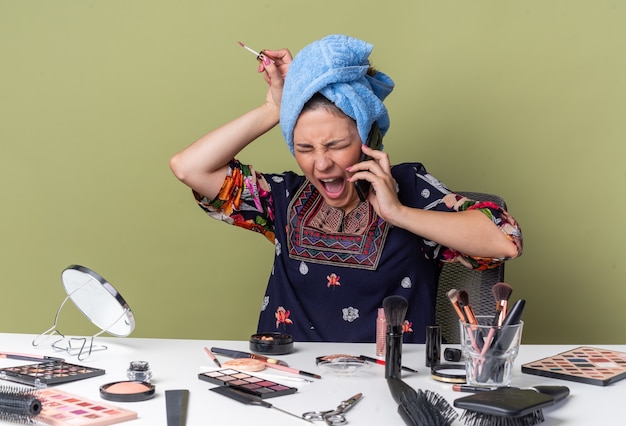 Chica morena joven enojada con el pelo envuelto en una toalla sentada en la mesa con herramientas de maquillaje gritando a alguien en el teléfono y sosteniendo brillo de labios