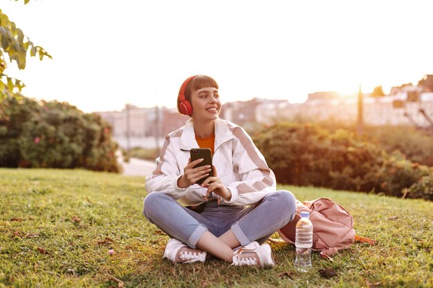 Chica morena joven feliz en jeans y chaqueta se sienta en la hierba y sostiene el teléfono inteligente Atractiva mujer de pelo corto escucha música en los auriculares