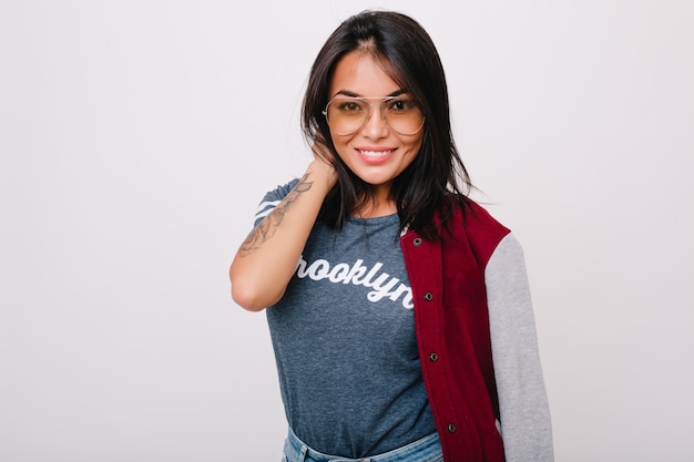 Chica morena ligeramente bronceada posando con sonrisa aislada. Modelo de mujer de pelo negro con gafas y bombardero divirtiéndose.