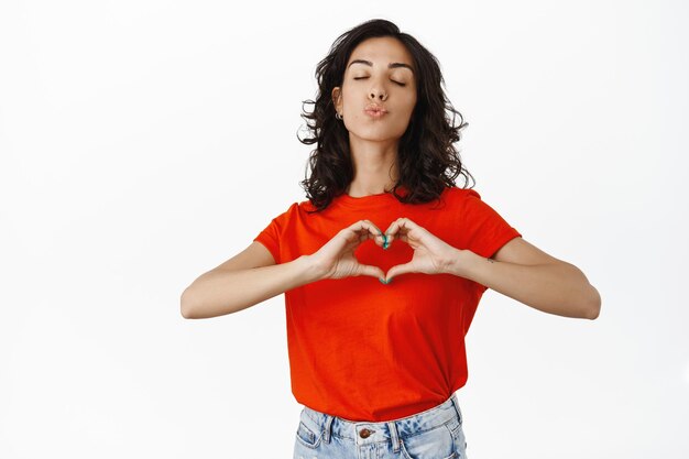 Chica morena linda y tonta, labios fruncidos y muestra el corazón Te amo firmar, besando a amante, haciendo confesión de amor, de pie sobre fondo blanco.