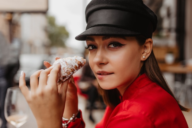 Chica morena se muerde el labio a la vista de moño con glaseado. Retrato de primer plano de blogger con estilo que viaja en Francia