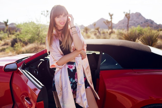 Chica morena muy seductora posando cerca de coche convertible deportivo moderno rojo.