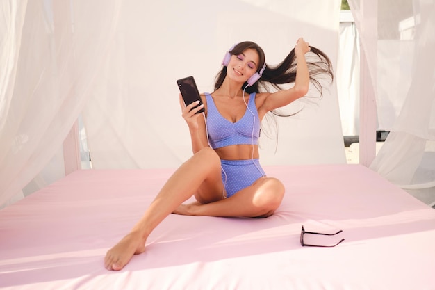 Chica morena muy sonriente en lindo traje de baño felizmente escuchando música en auriculares en la cama de playa con cortinas transparentes alrededor