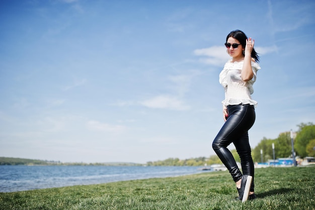 Chica morena en pantalones de cuero de mujer y gafas de sol de blusa blanca posadas sobre hierba verde contra la playa del lago