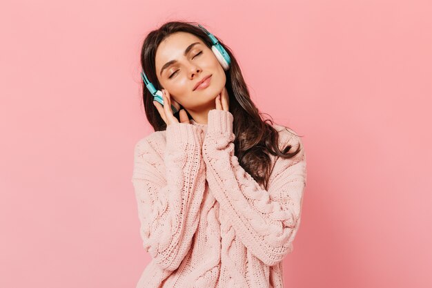 Chica morena con placer escucha música en auriculares. Mujer en traje rosa sonriendo sobre fondo aislado.