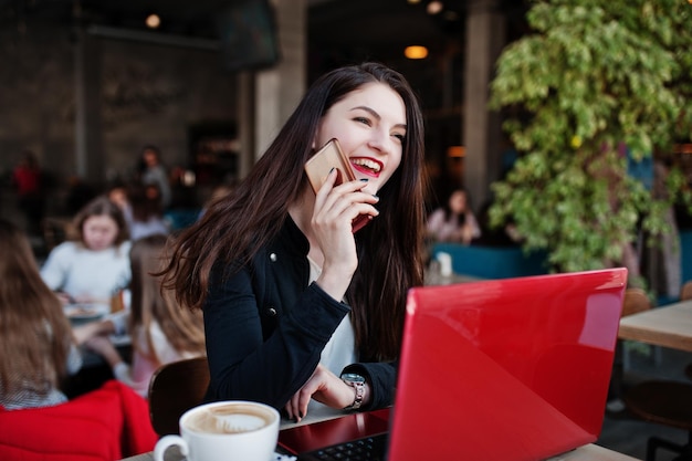 Chica morena sentada en un café trabajando con una laptop roja y hablando por teléfono móvil