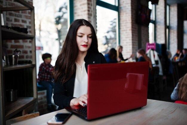 Chica morena sentada en un café y trabajando con una laptop roja