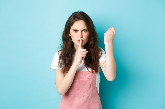 Chica morena seria y enojada agitando el puño y frunciendo el ceño con desaprobación, regañando el mal comportamiento de la persona, diciendo que se quede callado, sosteniendo el dedo en los labios, fondo azul.