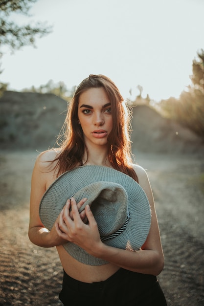 Chica morena sexy en la naturaleza que cubre su cuerpo con un sombrero con el sol brillando detrás de ella