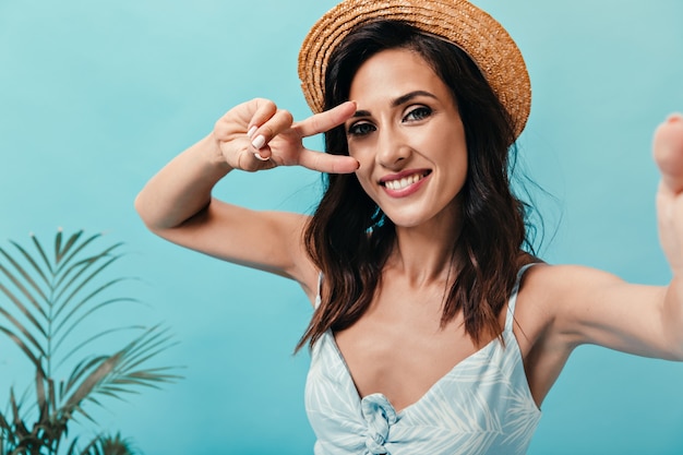 Chica morena con sombrero de paja muestra el signo de la paz y toma selfie sobre fondo azul. Señora adulta bonita en vestido ligero elegante hace la foto.